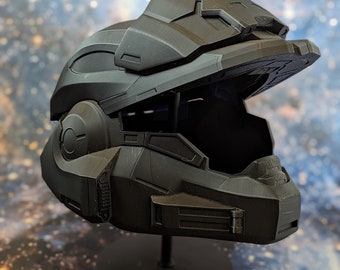 Könnte beinhalten: Ein schwarzer 3D-gedruckter Helm mit einem offenen Visier. Der Helm steht auf einem schwarzen Ständer. Der Hintergrund ist ein verschwommenes Bild einer Galaxie.