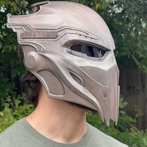 Könnte beinhalten: Ein metallisch-silberner Predator-Helm mit detailliertem Design. Der Helm hat ein Visier, aufwendige Schnitzereien und eine markante Kinnlinie. Das Gesamtdesign deutet auf ein futuristisches oder Science-Fiction-Thema hin.