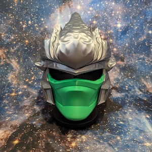 Op de afbeelding: Een zilveren en groene 3D-geprinte masker met een stekelig ontwerp. Het masker heeft een groene gezichtsbedekking en een zilveren helm met een puntige bovenkant.