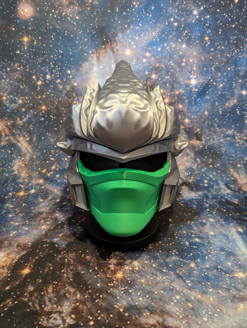 Op de afbeelding: Een zilveren en groene 3D-geprinte masker met een stekelig ontwerp. Het masker heeft een groene gezichtsbedekking en een zilveren helm met een puntige bovenkant.