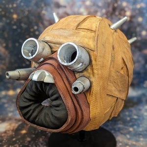 Peut inclure: Un masque de personnage Star Wars brun et argenté avec une surface texturée et une pièce buccale noire. Le masque est sur un support noir.