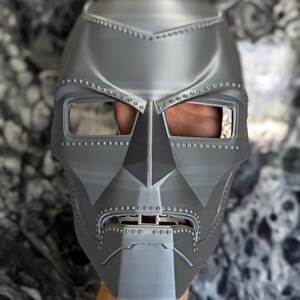 Può includere: Una maschera grigia stampata in 3D con un design dettagliato, che ricorda l'elmo di un supereroe. La maschera ha una parte superiore appuntita, due fori per gli occhi e un'apertura per la bocca.