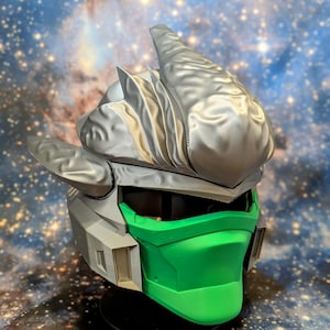 Op de afbeelding: Een 3D-geprinte helm met een zilveren en groene kleurstelling. De helm heeft een grote, puntige kam en een gezichtsmasker met een groene vizier.