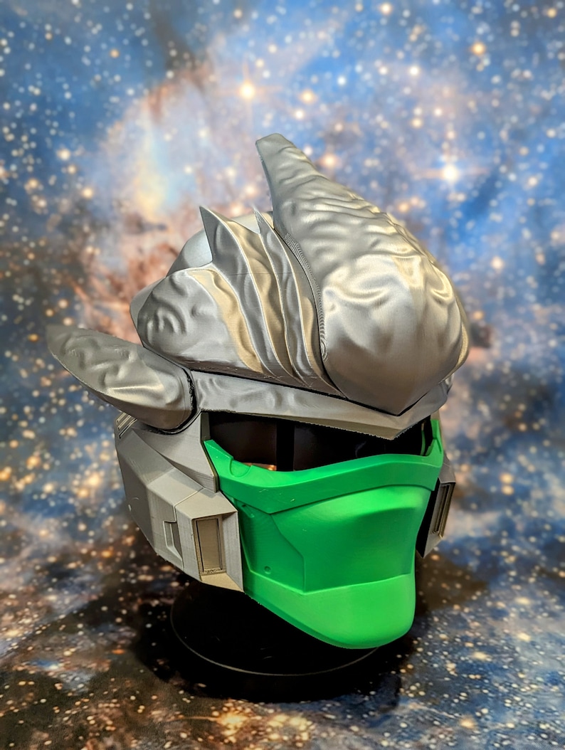 Op de afbeelding: Een 3D-geprinte helm met een zilveren en groene kleurstelling. De helm heeft een grote, puntige kam en een gezichtsmasker met een groene vizier.