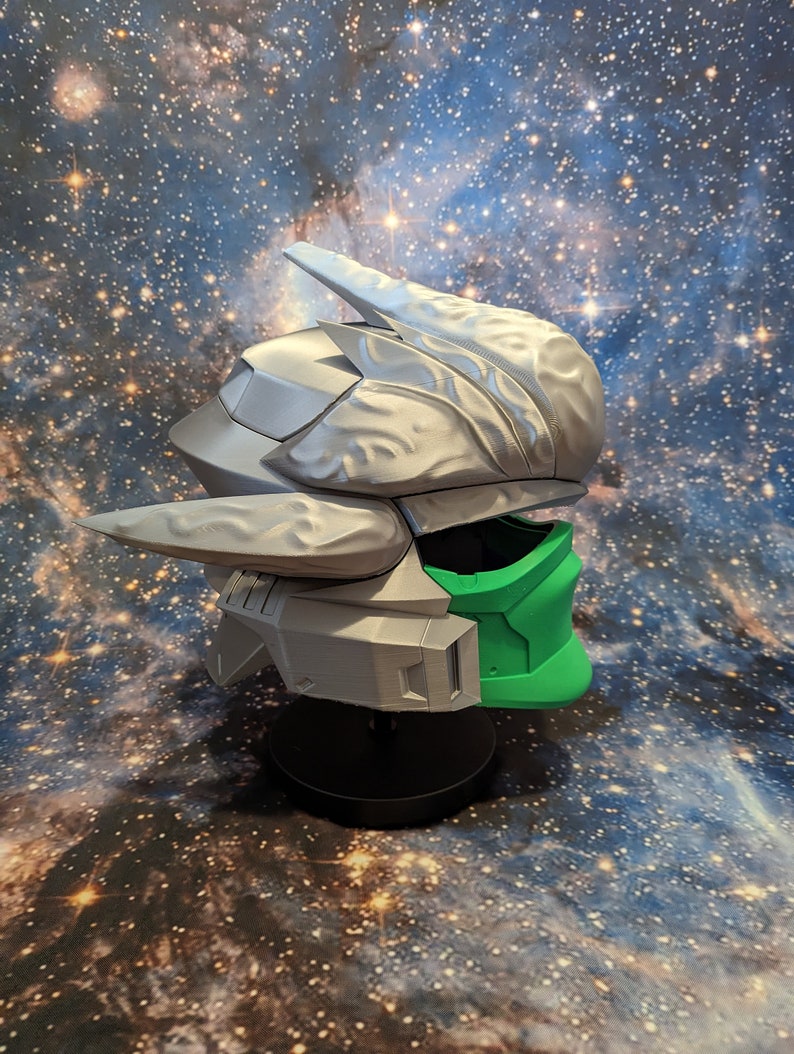 Op de afbeelding: Een 3D-geprinte helm met een zilveren en groene kleurstelling. De helm heeft een futuristisch ontwerp met ingewikkelde details en een vizier.
