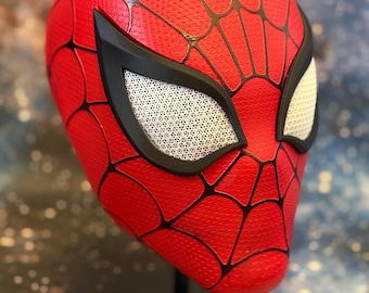 May include: A red Spider-Man mask with black webbing and black eye frames. The eye lenses are white with a mesh pattern. The mask is displayed on a black stand, showcasing the iconic superhero design.