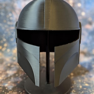 May include: A black plastic Mandalorian helmet replica on a black stand. The helmet has a visor and a textured surface.