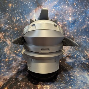 Op de afbeelding: Een 3D-geprint model van een futuristische helm met een grijze afwerking. De helm heeft een puntige bovenkant en een reeks richels en groeven.