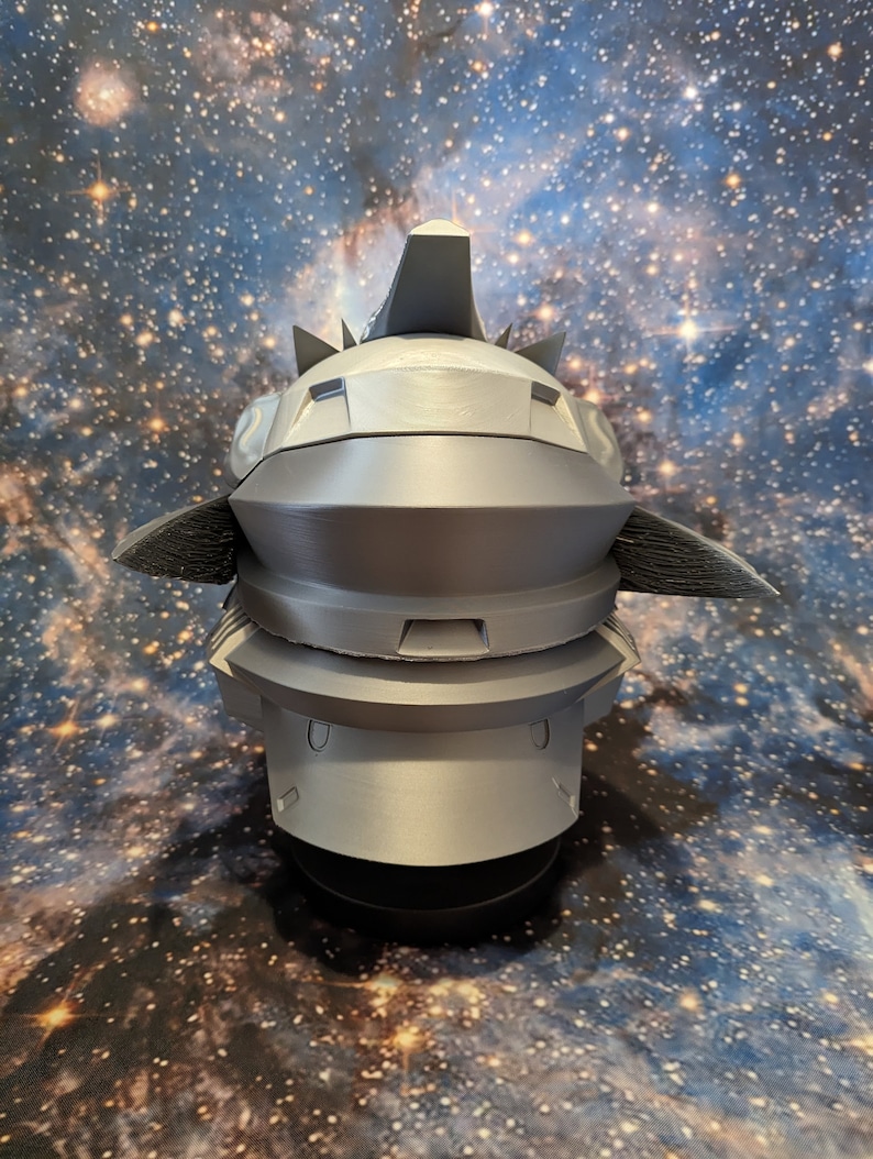 Op de afbeelding: Een 3D-geprint model van een futuristische helm met een grijze afwerking. De helm heeft een puntige bovenkant en een reeks richels en groeven.
