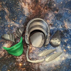 Op de afbeelding: Een set 3D-geprinte helmdelen voor cosplay of kostuum maken. De set bevat een groene vizier, een grijze helm en verschillende grijze onderdelen die lijken op hoorns of vleugels.