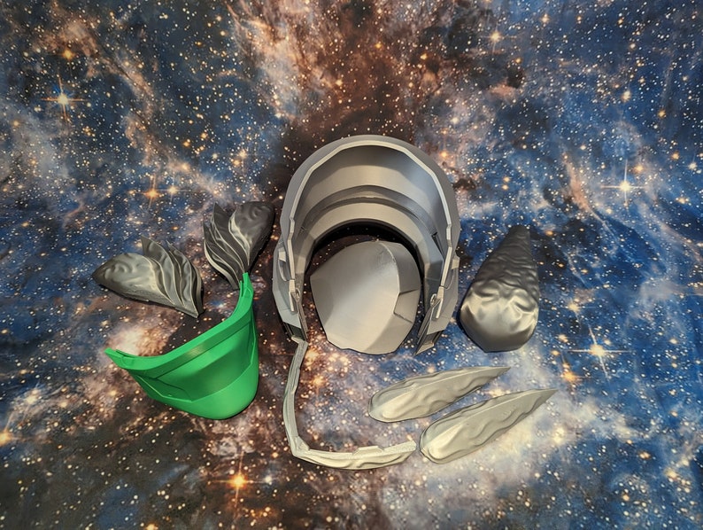 Op de afbeelding: Een set 3D-geprinte helmdelen voor cosplay of kostuum maken. De set bevat een groene vizier, een grijze helm en verschillende grijze onderdelen die lijken op hoorns of vleugels.