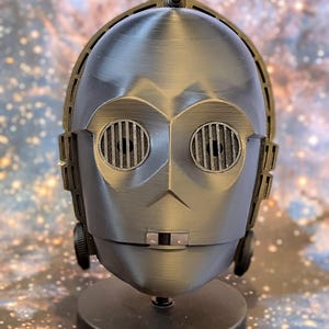 May include: A metallic gray C-3PO head replica, a character from the Star Wars franchise, is displayed on a black stand. The robot head features detailed facial features, including circular eye-like vents and a distinctive nose.