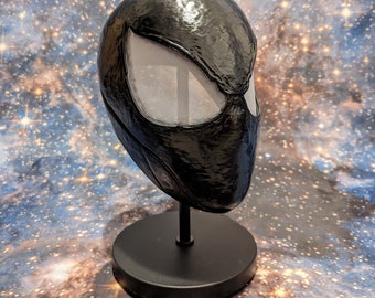 May include: A black and white Spider-Man mask with a mesh eye piece. The mask is on a black stand.