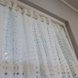 May include: A close-up of a white, crocheted curtain hanging in a window. The curtain has a delicate, openwork design with small, evenly spaced holes. The curtain is made of a soft, textured yarn and is partially covering the window.