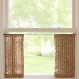 May include: A window with a white frame and two brown woven curtains. The curtains are rectangular and hang from wooden rods. The window looks out onto a green lawn and trees, with a white house in the background.
