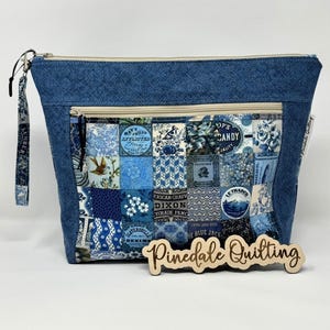 May include: A blue denim zippered pouch with a patchwork design featuring blue and white floral patterns and vintage-style text. The pouch has a zippered pocket on the front and a zippered closure. The pouch is labeled "Pinedale Quilting".