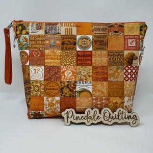 May include: A patchwork fabric zippered pouch with a brown handle. The fabric features a vintage design with a variety of labels and text in orange, yellow, and brown. The text includes "Fire Proof Stove Cream", "Orange", "Gold Mandarin Tea", and "Pinedale Quilting".