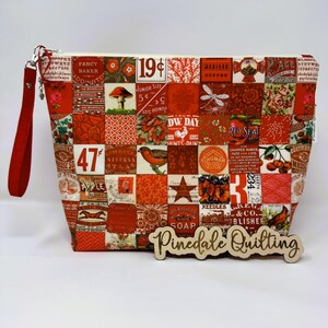 Puede incluir: Una bolsa de cremallera de tejido patchwork rojo y blanco con un asa roja. La bolsa tiene una variedad de diseños y textos de inspiración vintage, incluyendo "19¢", "Talla Junior", "Sello Rojo", "47¢", "Atlas Niversa", "DW. Day", "Kingman Co.", "Jabón Mágico Los Biegheles", "Agujas & Co. Blisher", y "Pinedale Quilting".