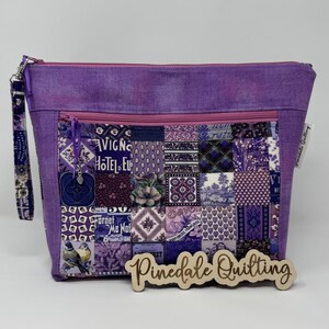 May include: A quilted purple zippered pouch with a wrist strap. The front features a patchwork design with various purple patterns, including floral, geometric, and text-based squares. A wooden sign that says "Pinedale Quilting" is in front of the pouch.