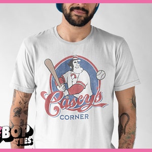 May include: White t-shirt with a vintage-style graphic of a cartoon baseball player holding a bat and a baseball. The graphic is in red, blue, and white and says "Casey's Corner".