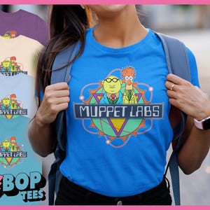 Może przedstawiać: Niebieska koszulka z logo Muppet Labs, zawierająca napis "Muppet Labs" w prostokątnym banerze. Projekt przedstawia postacie z kreskówek i geometryczne kształty. Widoczne są również inne koszulki w różnych kolorach.