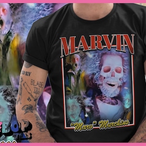Puede incluir: Camiseta negra con un gráfico de una calavera y el nombre "MARVIN" en rojo y blanco. La camiseta también incluye el texto "The Mara Marchins" e imágenes de un personaje con cara de calavera. El brazo está tatuado.
