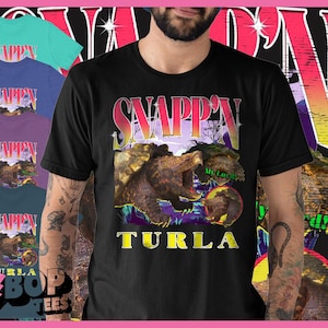 May include: Black t-shirt with a graphic of a snapping turtle and the word "TURTLA" in yellow. The word "SNAPP'N" is above the turtle graphic in a retro font. Other colored t-shirts are visible in the background.