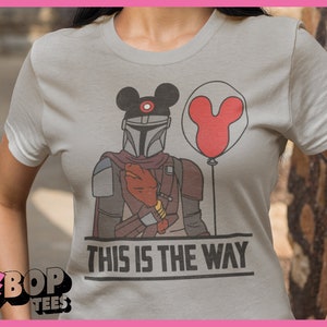 May include: A grey t-shirt featuring a graphic of a Mandalorian wearing a Mickey Mouse hat and holding a red Mickey Mouse balloon. The text "This is the way" is printed below the graphic.