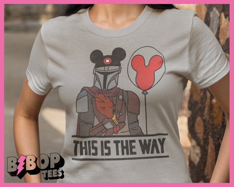 May include: A grey t-shirt featuring a graphic of a Mandalorian wearing a Mickey Mouse hat and holding a red Mickey Mouse balloon. The text "This is the way" is printed below the graphic.
