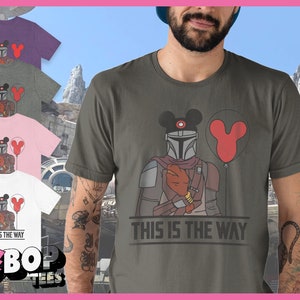 May include: A green t-shirt featuring a graphic of a Mandalorian character wearing a Mickey Mouse hat and holding a red balloon. The text "This is the way" is printed on the shirt.