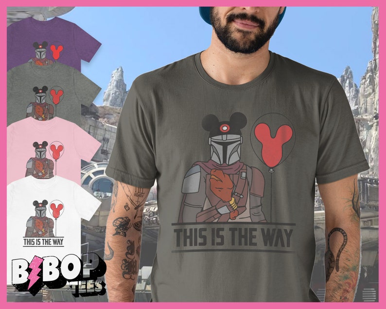 May include: A green t-shirt featuring a graphic of a Mandalorian character wearing a Mickey Mouse hat and holding a red balloon. The text "This is the way" is printed on the shirt.
