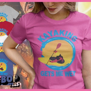 May include: A pink t-shirt with the words "KAYAKING GETS ME WET" in blue. The shirt features a graphic of a kayaker in a yellow and orange sunset. Other shirts in gray, red, white, and blue are visible.