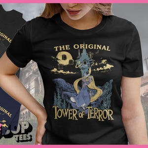 May include: A black short-sleeve t-shirt featuring a graphic print of a character with long blonde hair standing before a tall tower. The text reads THE ORIGINAL TOWER OF TERROR. The design includes a moon, clouds, and a mountainous landscape.