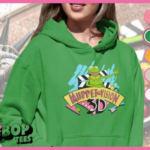Puede incluir: Sudadera con capucha verde con un gráfico de Muppet Vision 3D que presenta a Kermit la Rana frente a una pantalla de cine. El gráfico es de estilo retro con colores brillantes y un diseño de estrella.