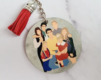 May include: A round keychain with a red tassel. The keychain features a cartoon illustration of a group of people, including a woman with long brown hair, a man with a yellow shirt, and a girl with red hair. The background is a light gray color.