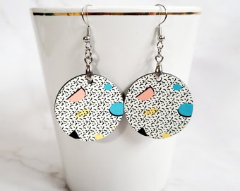 May include: A pair of round earrings with a white background and a black dotted pattern. The earrings feature colorful geometric shapes in pink, blue, yellow, and black. The earrings are hanging from silver-colored hooks.