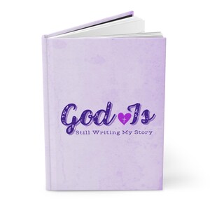 Pode incluir: Um diário de cor lavanda com o texto "God Is" em letras roxas com um pequeno coração roxo contendo uma cruz branca. O texto "Still Writing My Story" está abaixo do texto principal.