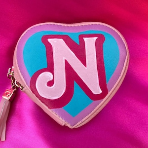 May include: A pink heart-shaped coin purse with a pink tassel. The purse has a blue, pink, and purple design with a white letter "N" in the center.