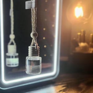 May include: A clear glass car air freshener bottle with a silver cap and a beige string. The bottle has a label that reads "90's KISS Car Diffuser". The air freshener is hanging in front of a glowing white-edged mirror.