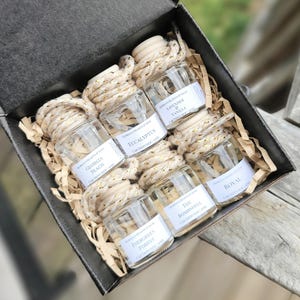 May include: A gift box containing six small glass car air fresheners. Each bottle has a wooden cap and a decorative gold and white rope. The labels read: Georgia Peach, Eucalyptus, Lavender & Vanilla, Evergreen Forest, The Bombshell, and Royal.