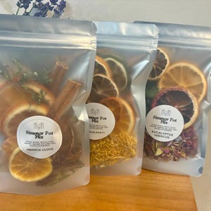 May include: Three clear pouches filled with dried orange slices, cinnamon sticks, and herbs. Each pouch has a white label with the text "Simmer Pot Mix". The pouches are on a wooden surface.