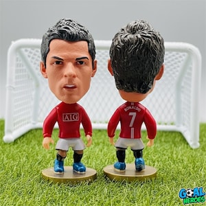 Peut inclure: Deux figurines à tête branlante d'un joueur de football en maillot rouge avec le numéro 7. La figurine de face a un visage détaillé et le mot "AIG" sur le maillot. L'autre figurine montre le dos du maillot avec le nom "Ronaldo".