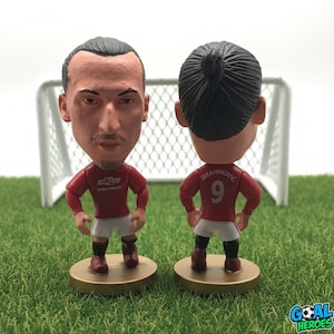 Peut inclure: Deux figurines bobblehead d'un joueur de football en maillot rouge et short blanc. L'une montre le visage du joueur, l'autre le dos du maillot avec "IBRAHIMOVIC" et le numéro "9". Les figurines sont sur une base dorée.