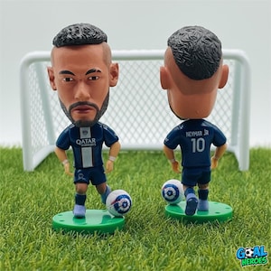 Könnte beinhalten: Zwei Neymar Jr. Wackelkopf-Figuren in Fußballuniform, die auf grünen Sockeln mit Fußbällen stehen. Die Figuren stehen vor einem kleinen weißen Fußballtor auf einer grünen Oberfläche. Die Wackelkopf-Figuren tragen den Text "Qatar Airways" auf der Vorderseite.