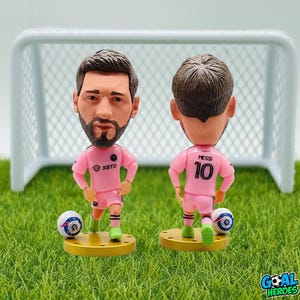 Puede incluir: Dos figuras de futbolista cabezonas, una de frente y otra de espaldas, con uniforme rosa. Las figuras están sobre una base dorada con un balón de fútbol. La camiseta del jugador tiene el número 10. Una pequeña portería en el fondo.