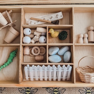 Pu&ograve; includere: Set di giocattoli in legno con vari elementi, tra cui una carriola, vasi, perline, un'anatra, pietre, una recinzione, un cestino e figure di legno. Il set &egrave; disposto in una scatola di legno con pi&ugrave; scomparti, perfetto per il gioco fantasioso.