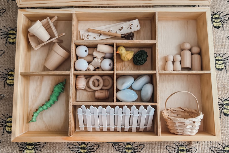 Pu&ograve; includere: Set di giocattoli in legno con vari elementi, tra cui una carriola, vasi, perline, un'anatra, pietre, una recinzione, un cestino e figure di legno. Il set &egrave; disposto in una scatola di legno con pi&ugrave; scomparti, perfetto per il gioco fantasioso.