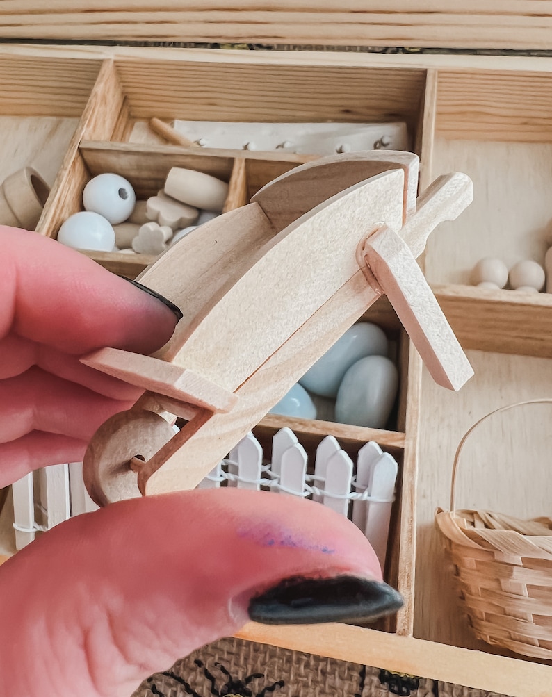 Pu&ograve; includere: Una carriola in legno in miniatura, parte di un set artigianale. La carriola &egrave; in legno di colore chiaro e ha una sola ruota. Altri pezzi artigianali in legno, tra cui perline e pezzi di recinzione, sono visibili sullo sfondo. Il set &egrave; in una scatola di legno.