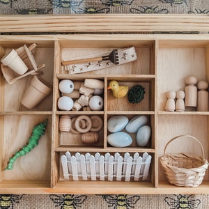 Pu&ograve; includere: Una scatola di legno con scomparti pieni di giocattoli in miniatura. Include piccoli vasi, una carriola, palline di legno, un'anatra, un bruco verde, figure di legno, una staccionata bianca, un cestino e altre forme di legno.