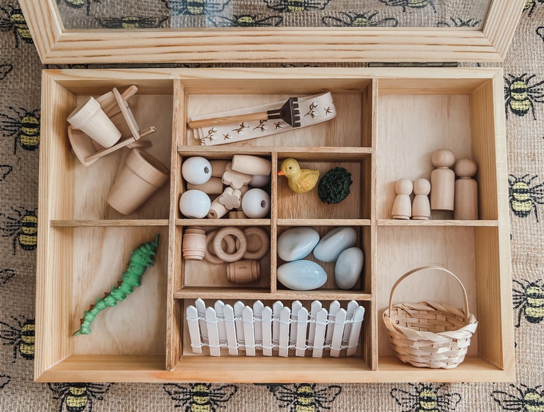 Pu&ograve; includere: Una scatola di legno con scomparti pieni di giocattoli in miniatura. Include piccoli vasi, una carriola, palline di legno, un'anatra, un bruco verde, figure di legno, una staccionata bianca, un cestino e altre forme di legno.